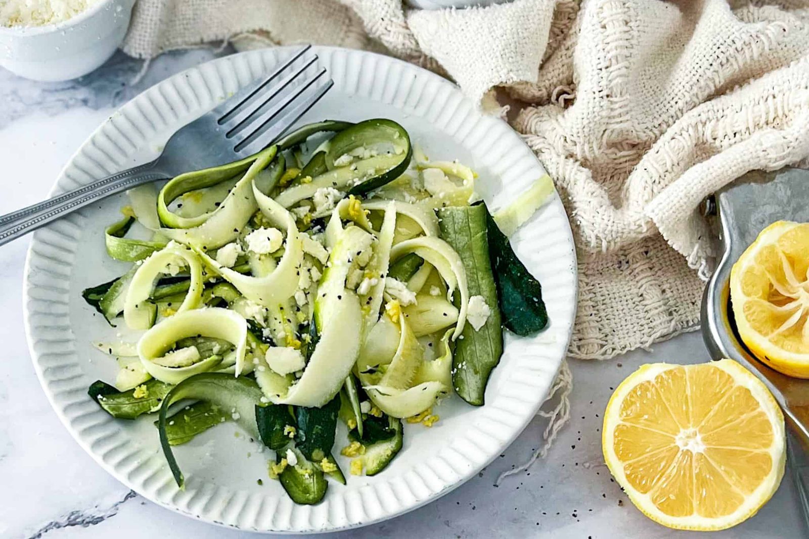 Raw courgette ribbons with feta on a white plate, with a fork, lemon halves and a bowl of feta