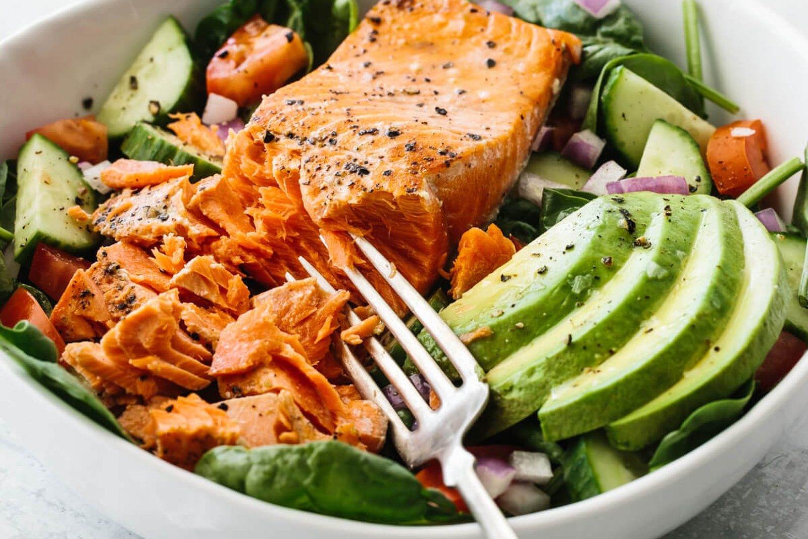 Seared salmon fillet flaked with a fork over spinach, with sliced avocado, cucumber and red onion in a white bowl