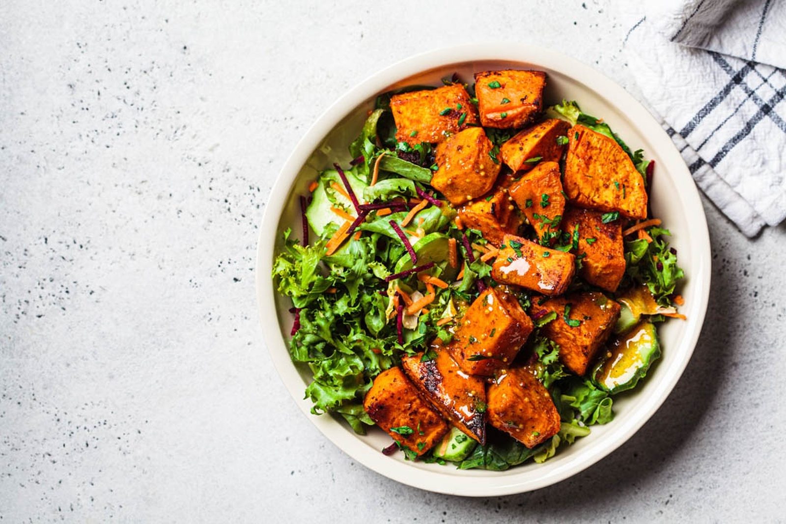 Roast sweet potato cubes on green salad leaves, avocado and grated carrot in a white bowl