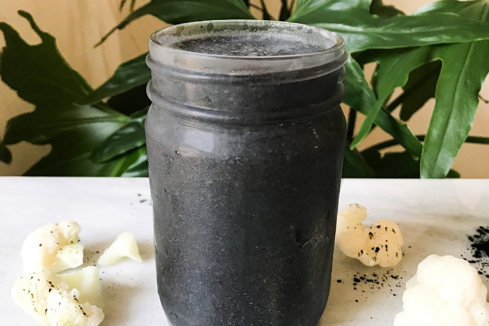 Black smoothie in a glass jar on a marble board with cauliflower florets and charcoal powder, in front of a green plant